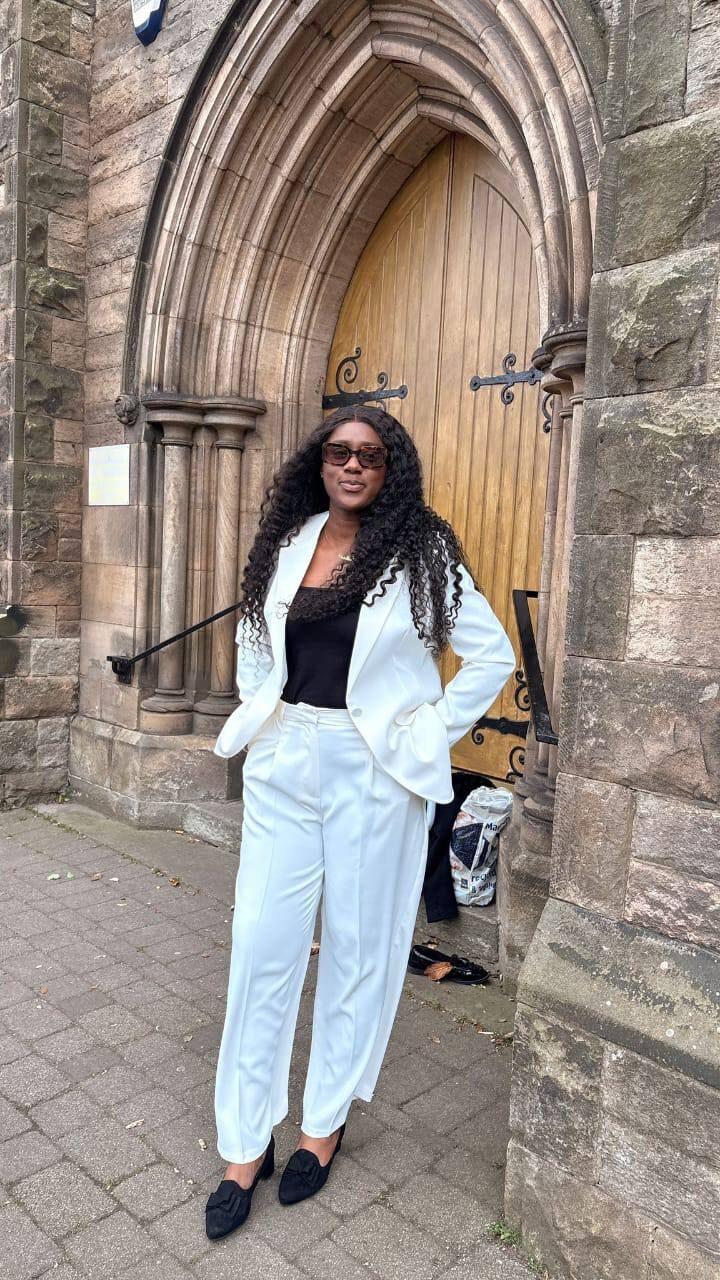 Omowunmi Esther Oyeleke standing before an arched stone doorway, in a white tailored suit